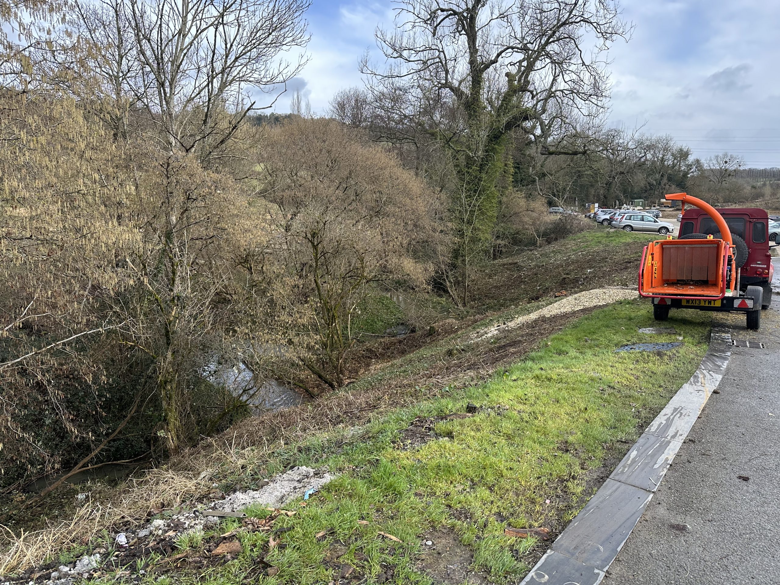 Wood chipper and utility vehicle parked beside the River Chelt embankment during clearance work