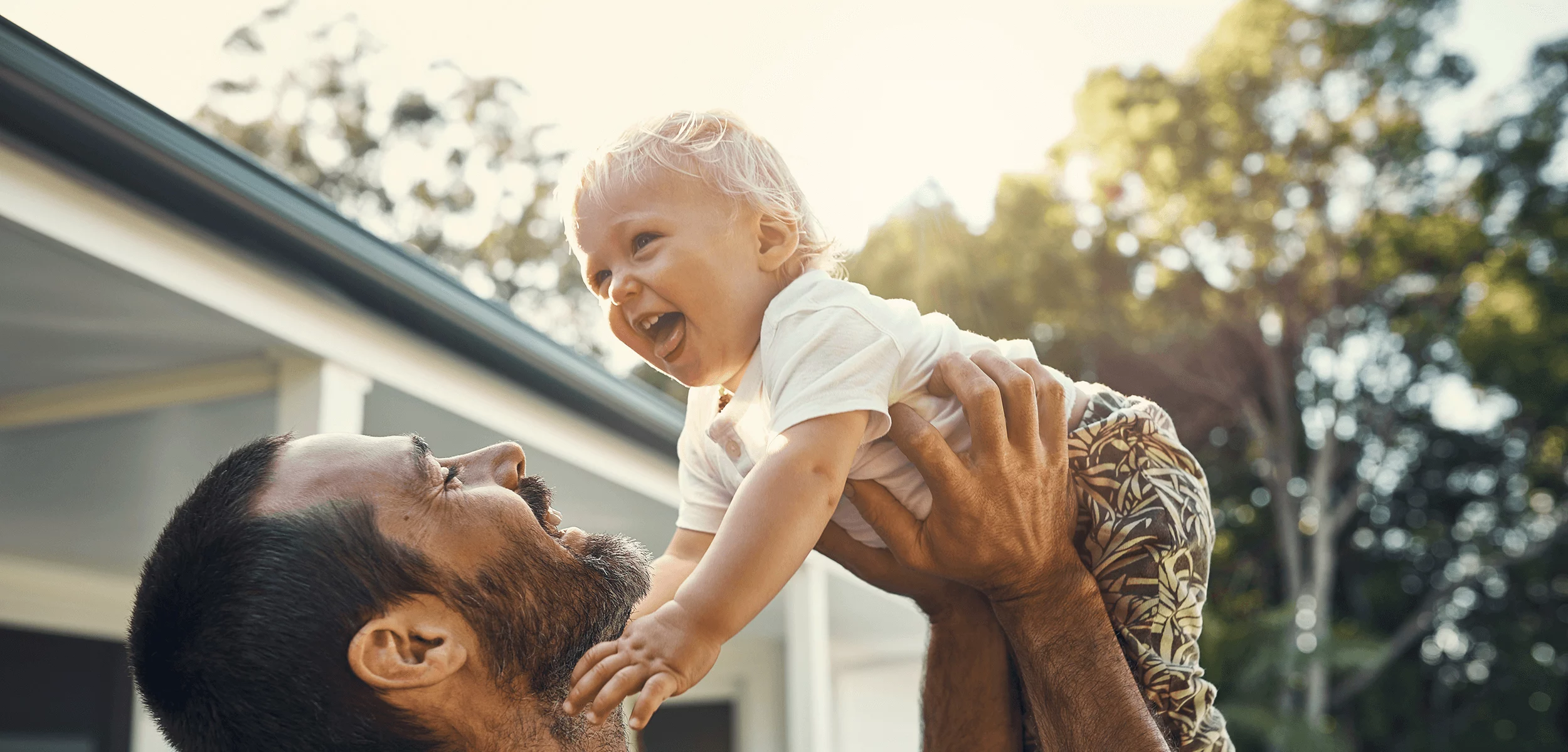 Parent lifting a smiling toddler outdoors in warm sunlight