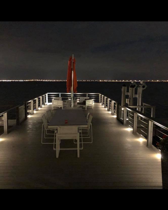 Modern waterfront deck at night with built-in floor lighting and outdoor dining area overlooking calm water