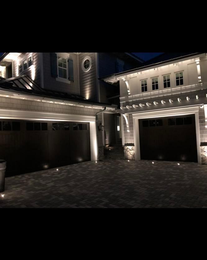 Upscale home exterior at night with warm LED accent lights outlining eaves, columns, and paver driveway