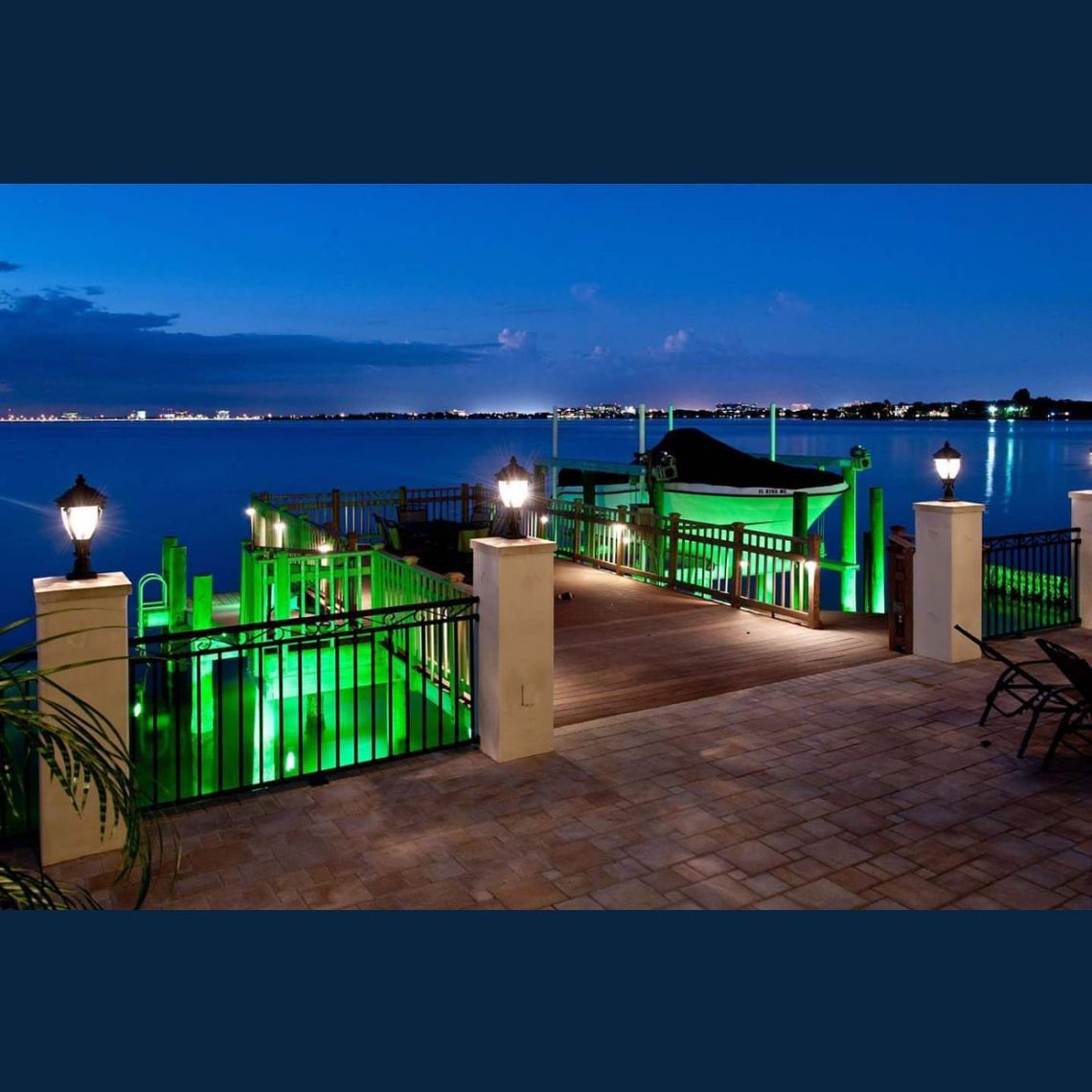 Waterfront dock at dusk with green accent lighting along pilings and a covered boat lift overlooking calm bay water