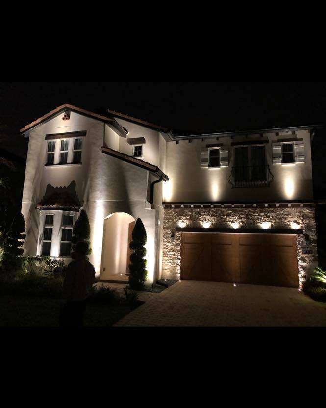 Two-story stucco home at night lit by warm architectural uplighting along walls and entryway