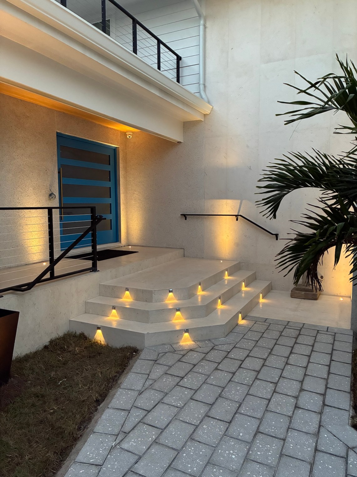 Modern residential entryway with recessed step lights, blue contemporary front door, and warm uplighting on pale concrete walls