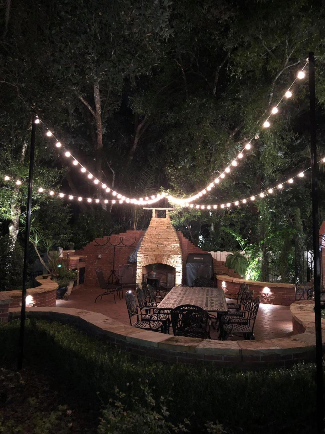 Outdoor patio dining area at night with string lights overhead and warm uplighting around a brick fireplace